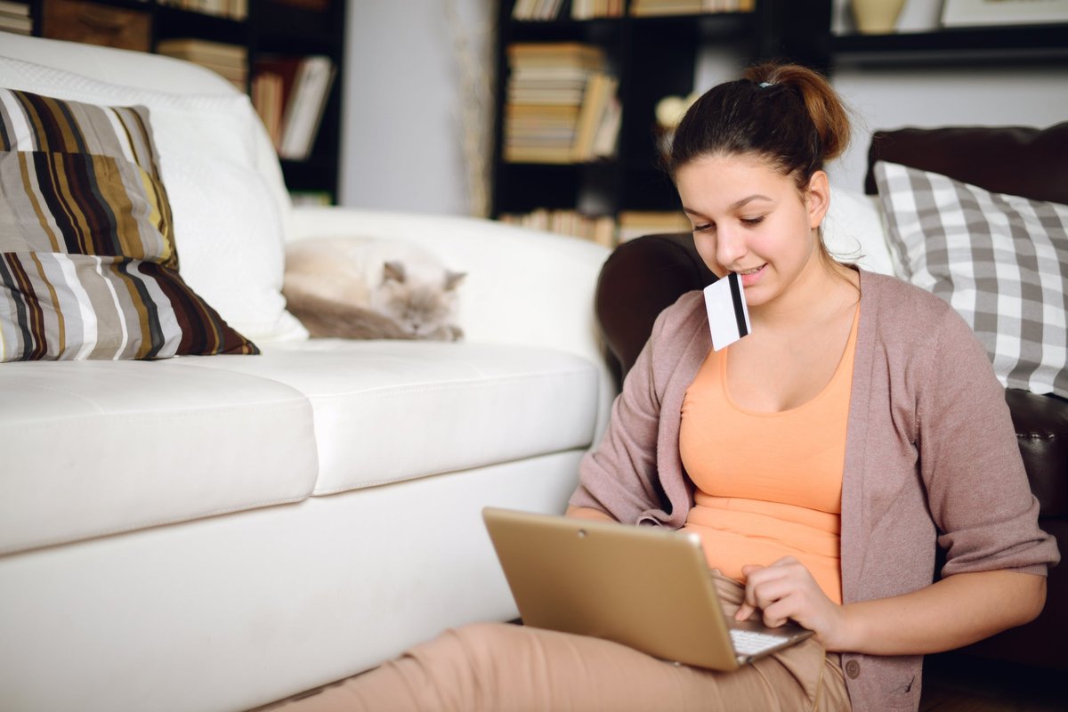 A young woman shops on her laptop holding her credit card while her cat lounges in the background