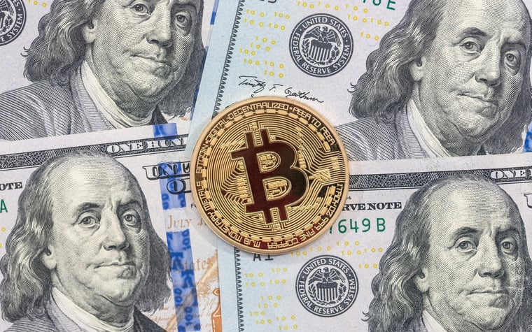 A physical gold coin marked with the Bitcoin B lying atop hundred dollar bills.