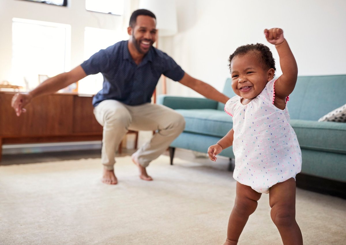 Baby fist-pumping and dad smiling with his arms open in the background