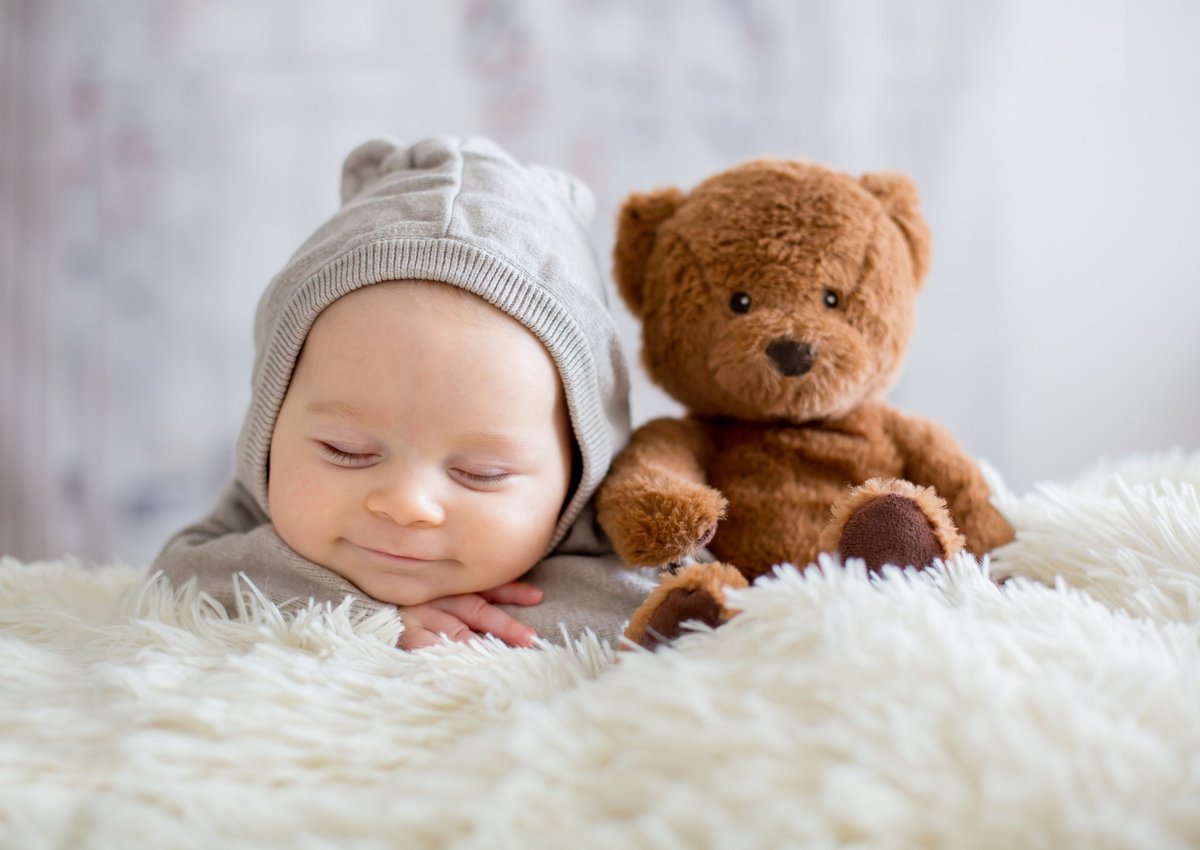 Sleepy, smiling baby in a knit hat next to a teddy bear on a fuzzy blanket.