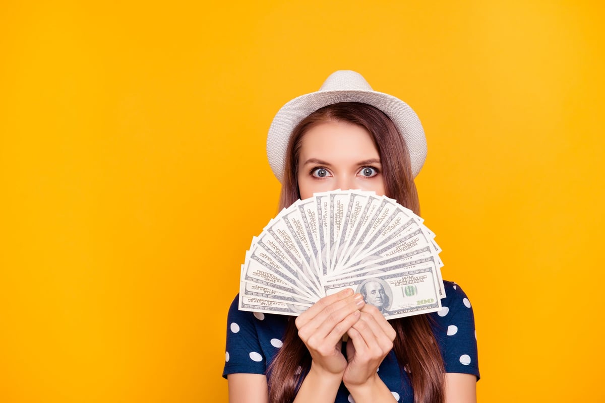 A young woman wearing a hat holds a fan of hundred dollar bills.