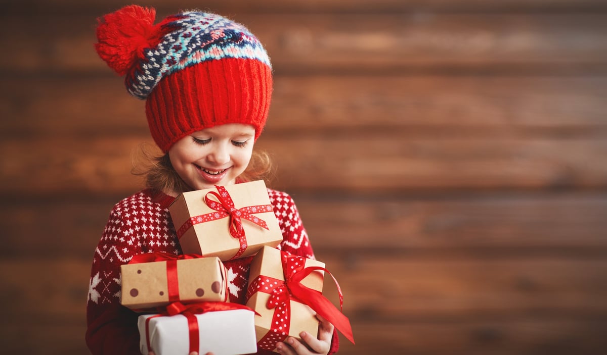 A grinning little girl in a knitted hat holds a lot of presents.