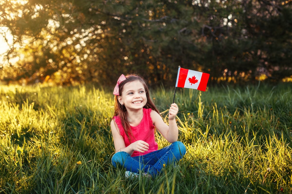 A child sits cross-legged in a meadow, smiling and holding a Canadian flag.