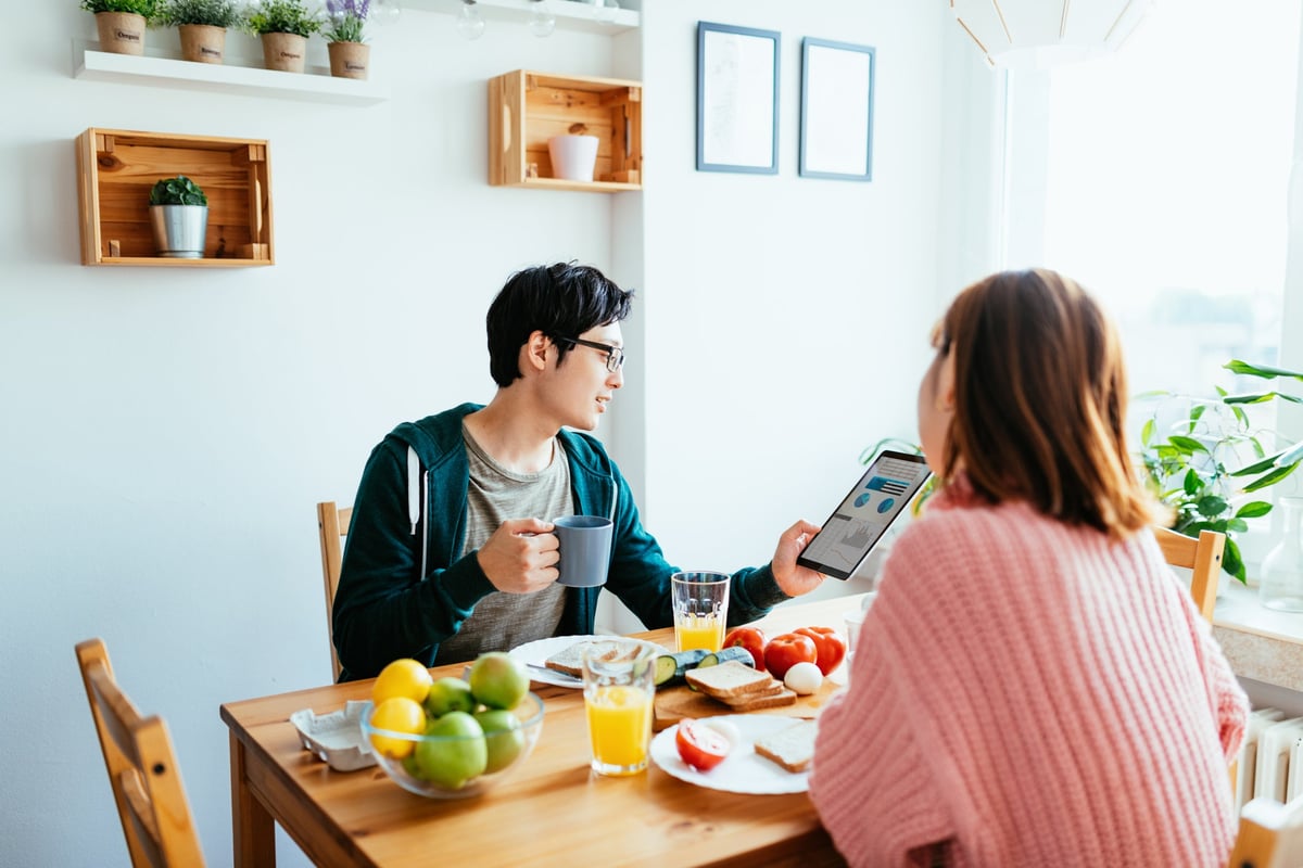 A couple eats breaksfast in their kitchen while the man looks at his tablet.