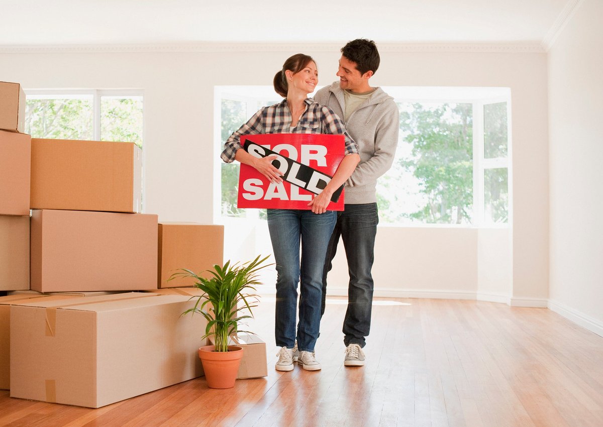 Couple holding sold real estate sign next to moving boxes.