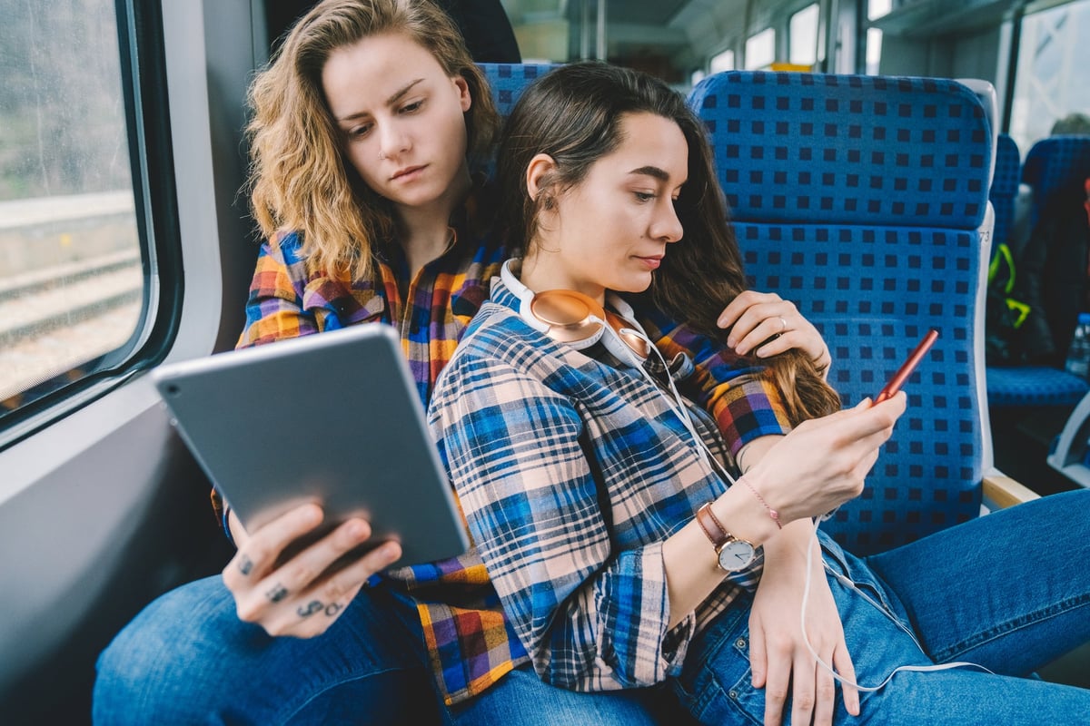 A couple sit together on a train. One looks at her tablet while the other looks at her phone.