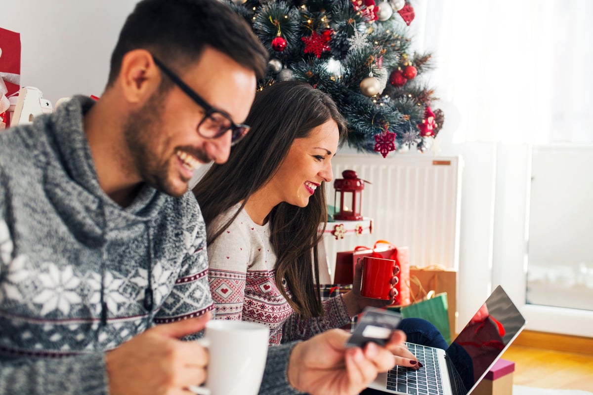 A smiling couple online shopping next to a Christmas tree.