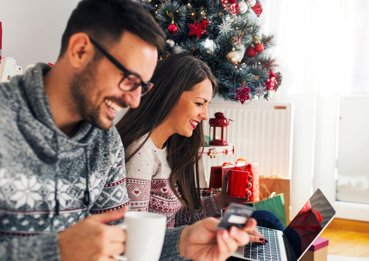 A smiling couple online shopping next to a Christmas tree.