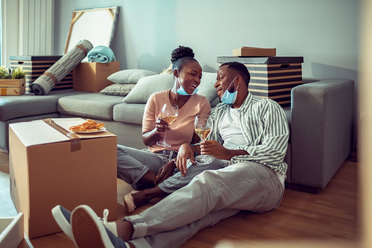 A couple sit on the floor of their new home, sipping wine and eating pizza.