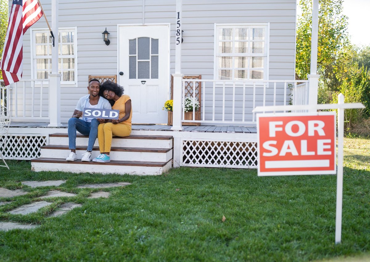 Couple sits on porch steps holding a house Sold sign.