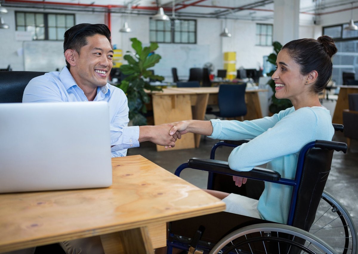 An employer shakes hands with a smiling applicant who is sitting in a wheelchair.