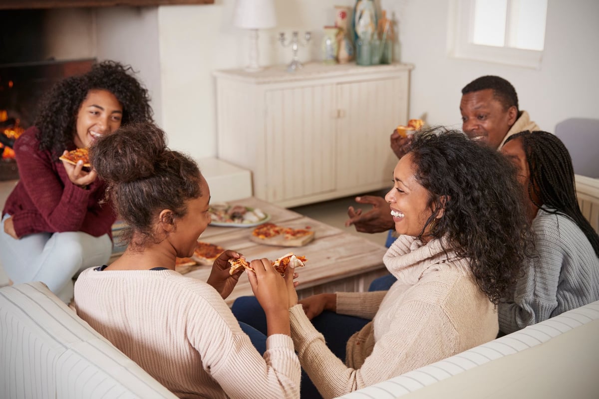 A family of five laughing together and eating pizza while sitting on the couch in their living room.