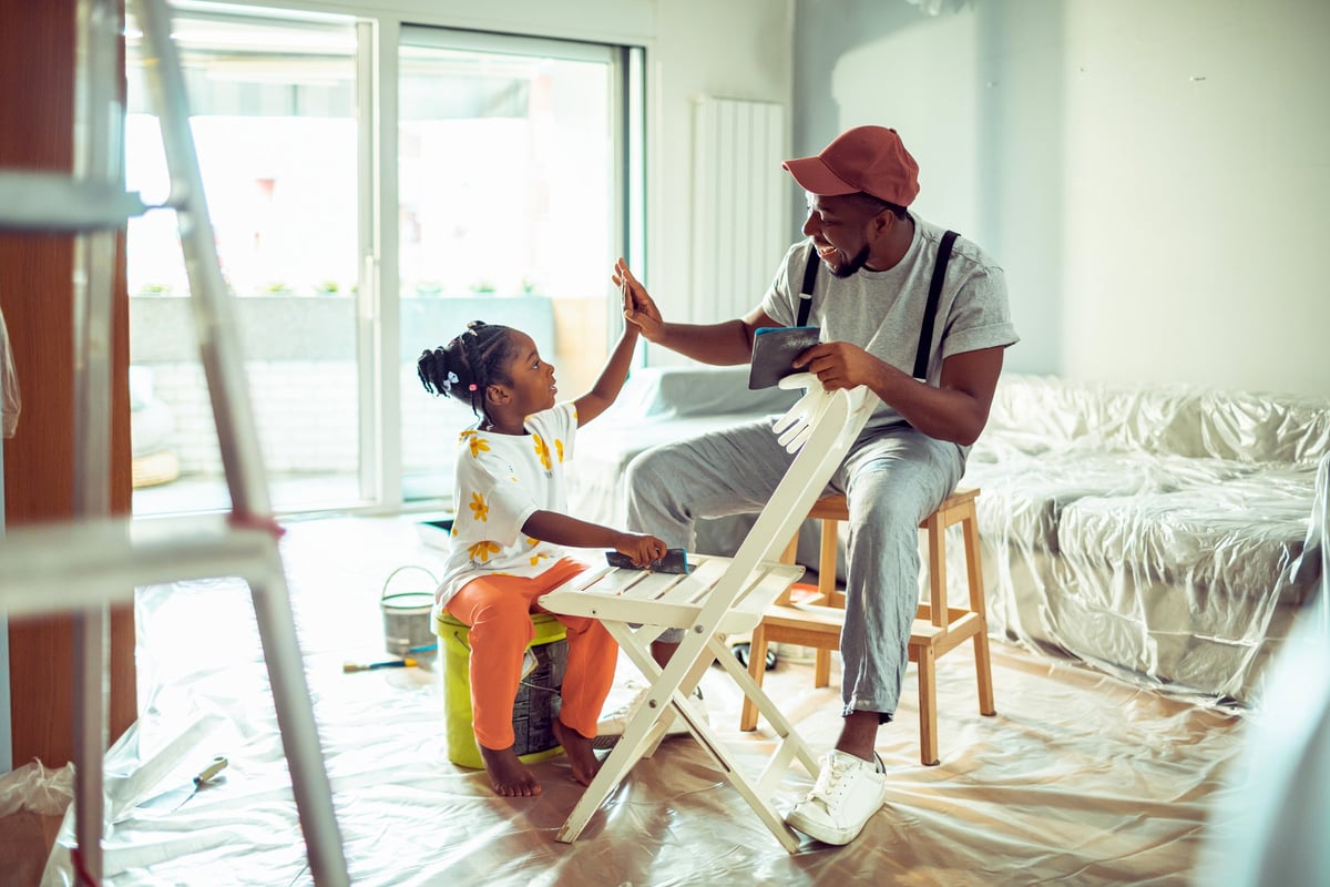 A father sits and high-fives his young daughter in the living room they are painting.