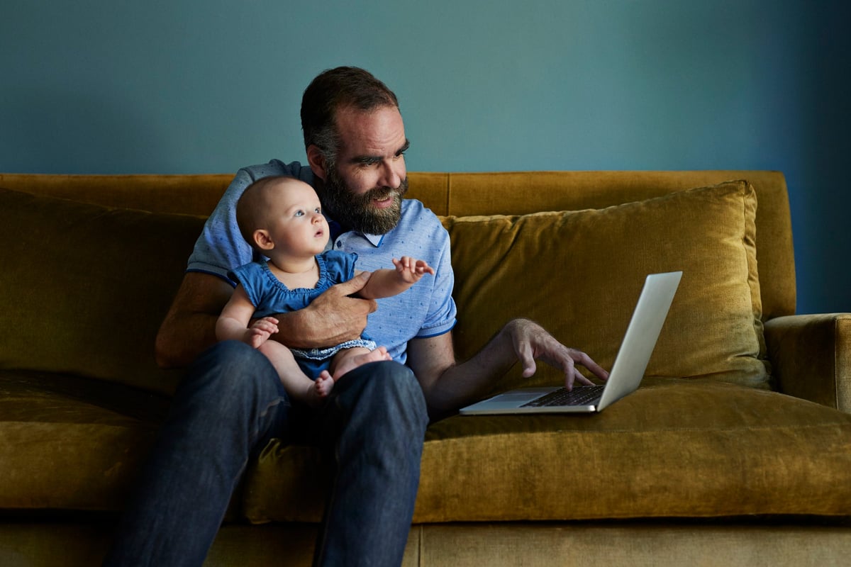 A father sits on a sofa with his baby while working on his laptop.
