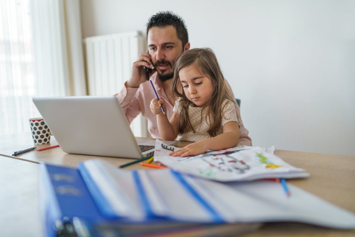 Father using laptop while on cell phone with young daughter coloring in his lap