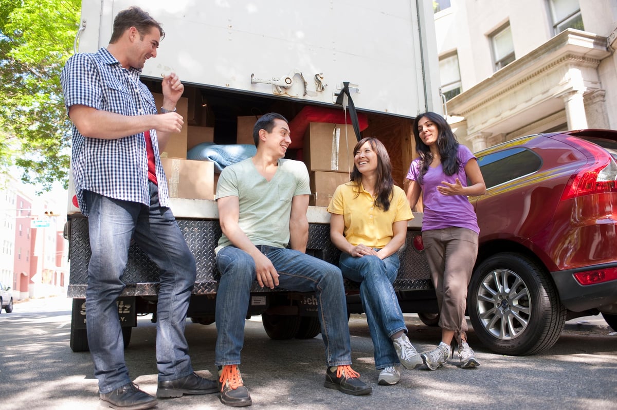 Four adults hanging out by loaded moving truck and car.