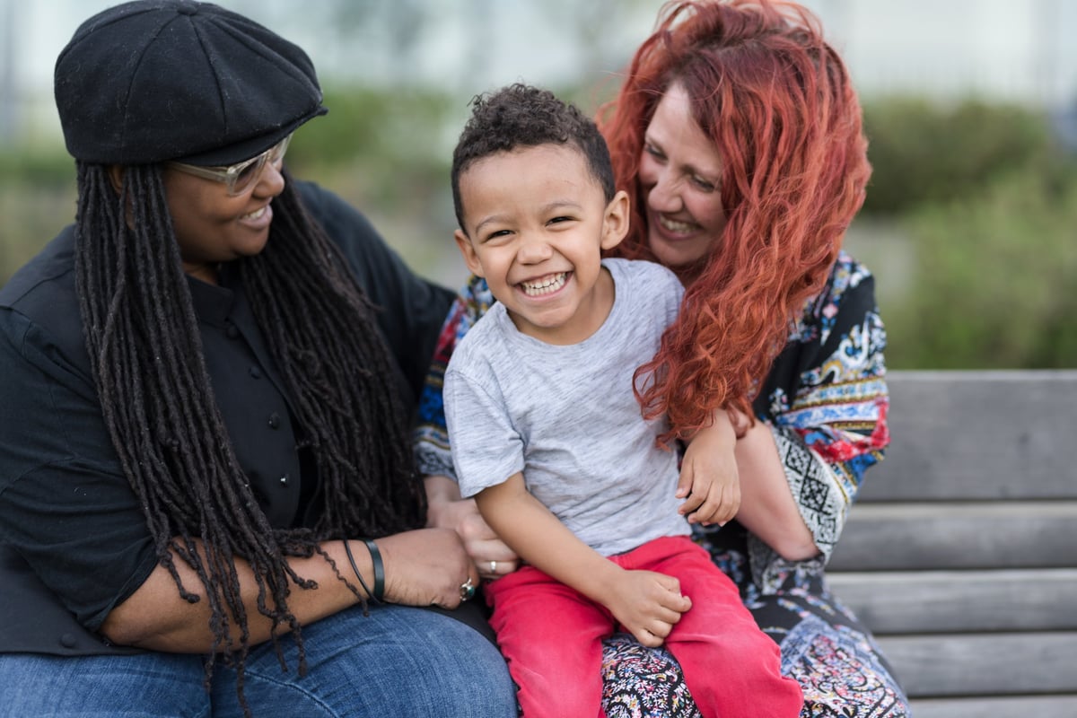 Two parents hold their child on their laps while sitting on a park bench and smiling.