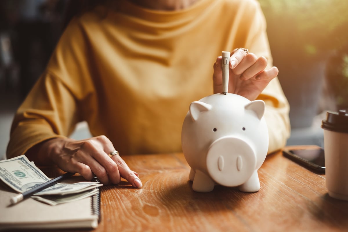 Person in a yellow sweater sitting behind desk and sliding a dollar bill into a piggy bank.