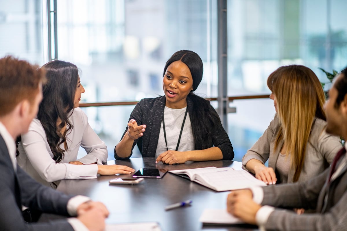 Young woman sitting at the head of a conference table leading a discussion with several co-workers.