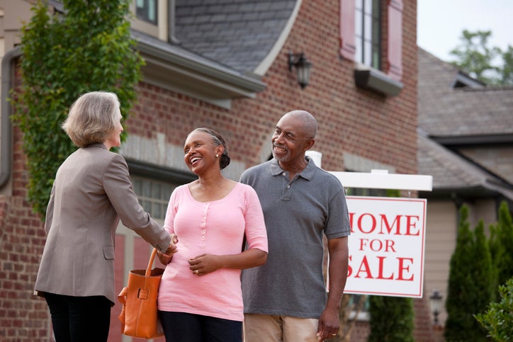A realtor shakes hands with a senior couple in front of a For Sale sign and house.