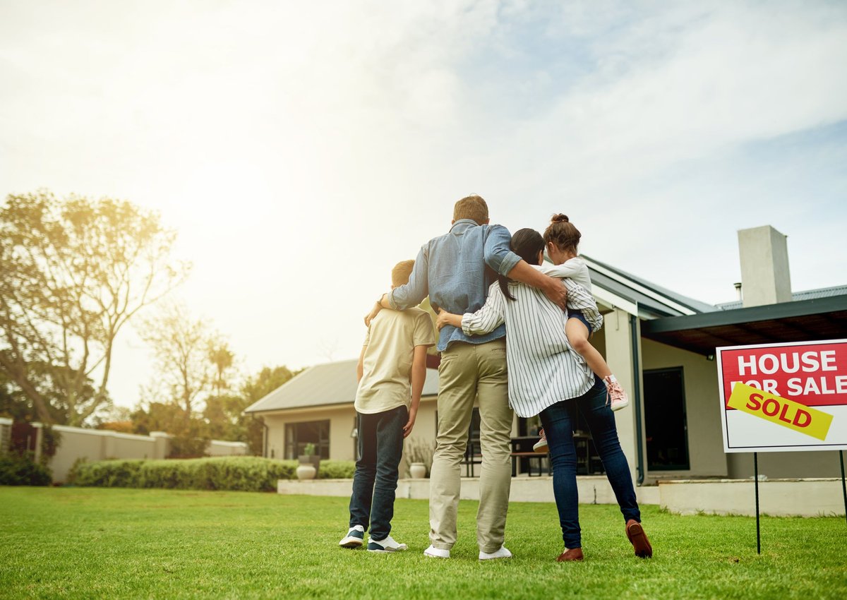 A family of four, arm in arm, look at a house with a "Sold" sign.