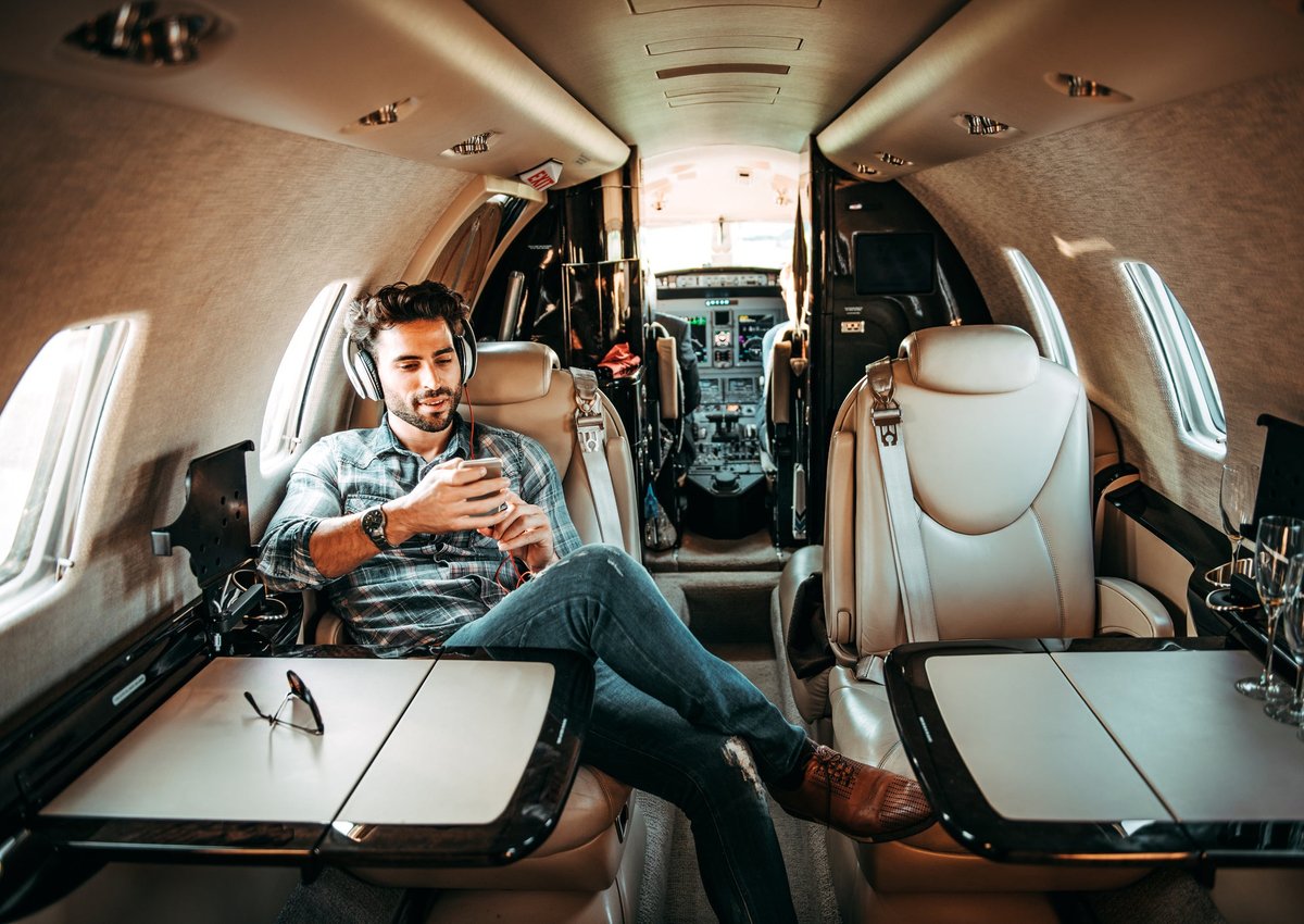 A person sitting in a private jet uses a smartphone and headphones while smiling.
