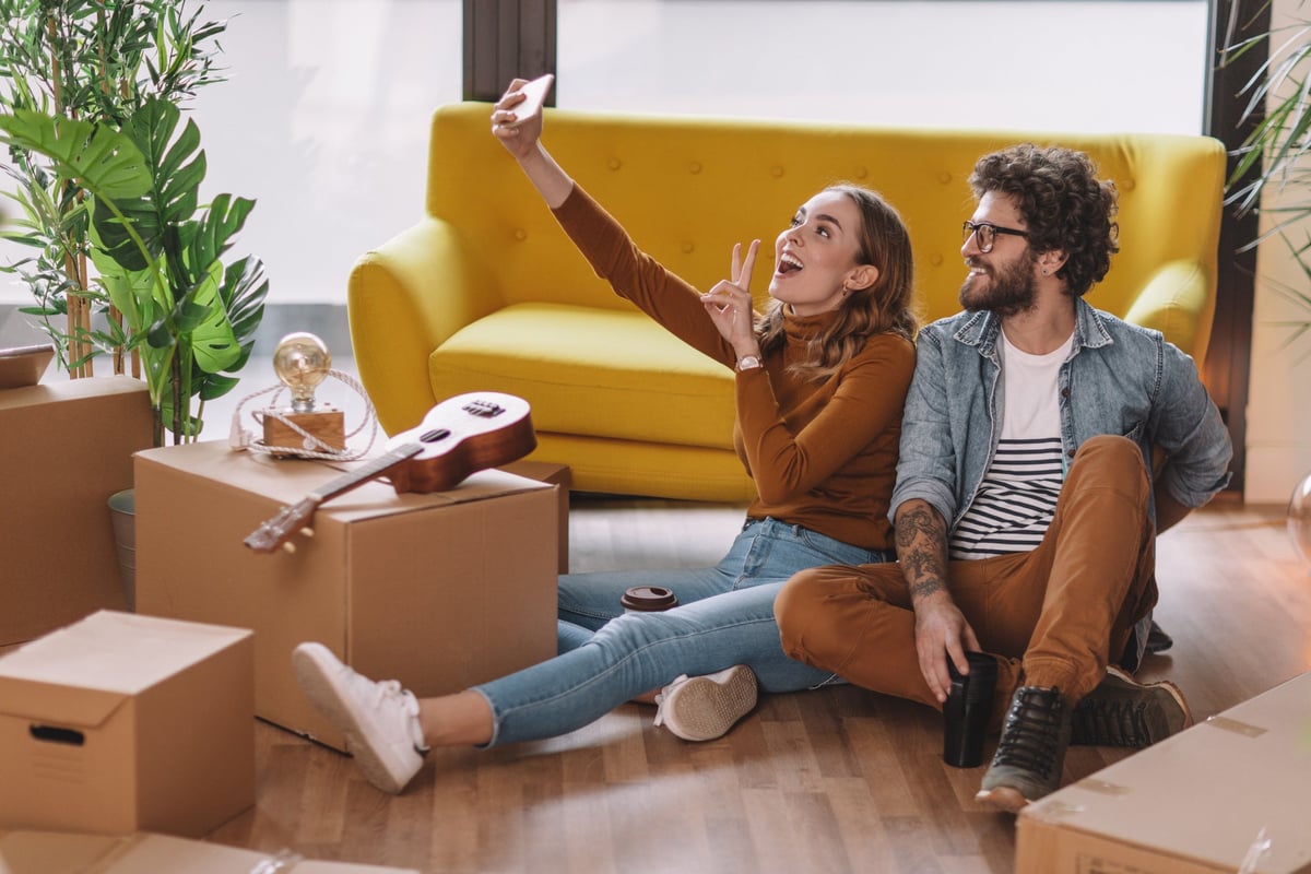 Two people take a selfie together while sitting on the floor next to moving boxes, a couch, and a ukulele.