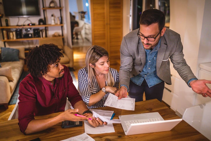 Three people discuss finances while filling out paperwork in their home.