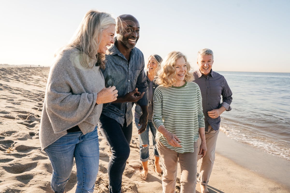 A group of people, some with gray hair, smile and converse as they walk along the beach.