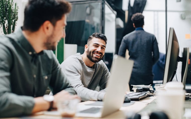 Bearded colleagues laughing together while sitting at desk in office.