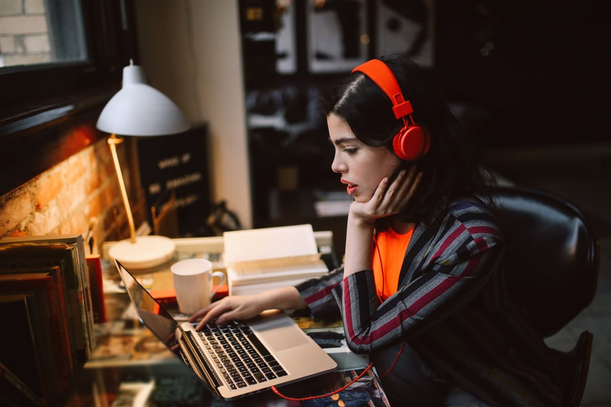 A person sits at a desk at home looking at a laptop screen and wearing headphones.