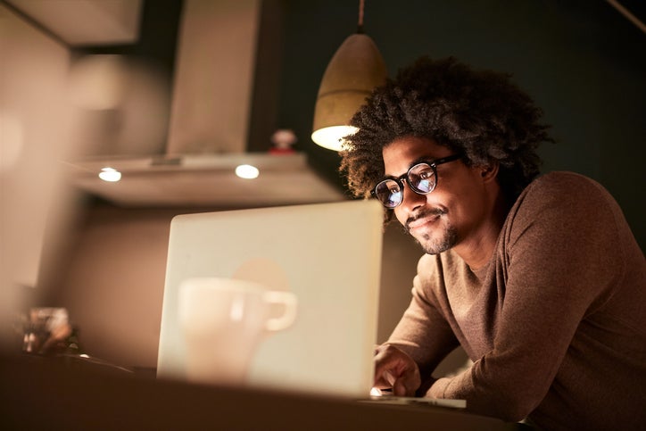 A man stands in a kitchen illuminated by the glow of a laptop screen.