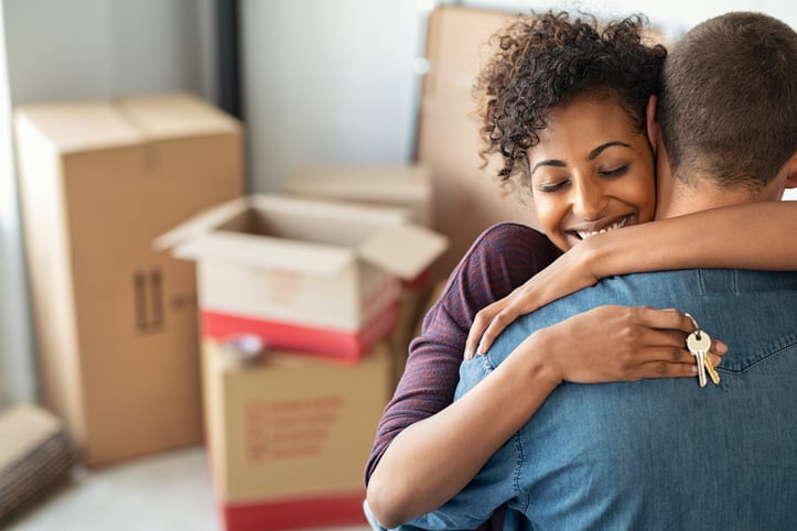 Couple holding keys to new house embrace in foreground, boxes in background.