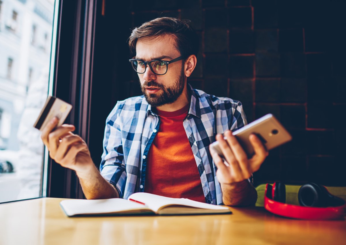 Man in blue checked shirt looks thoughtful with phone in one hand and credit card in the other.