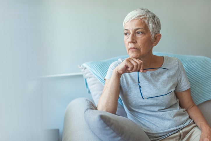 Grey-haired woman holding glasses rests hand on chin and looks thoughtful.