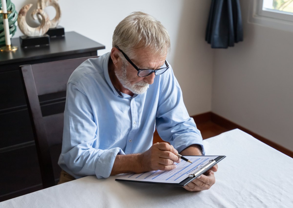 Grey-haired man sits at table and fills in form on clipboard.