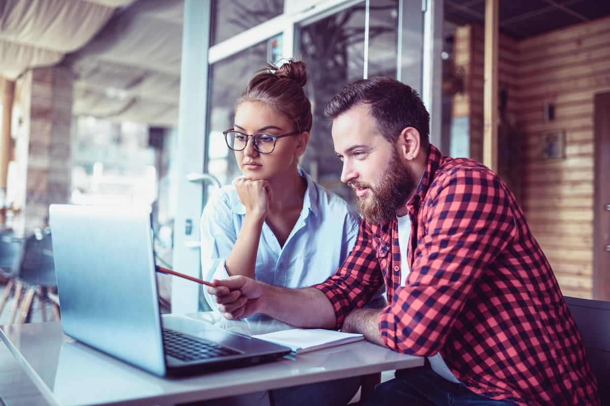 Two people in a casual public space discuss financial documents on a laptop and notepad.