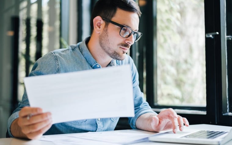 Man at desk holding document in one hand and typing with the other.