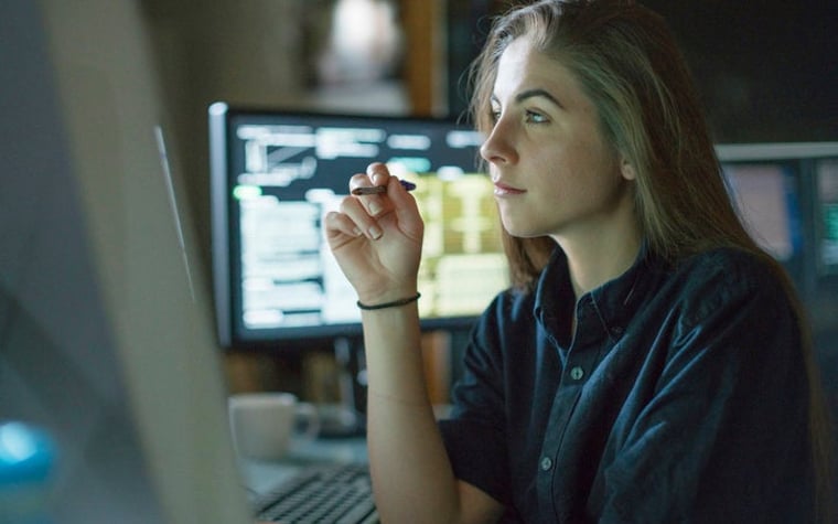 A young woman sits at a desk surrounded by monitors displaying financial data.
