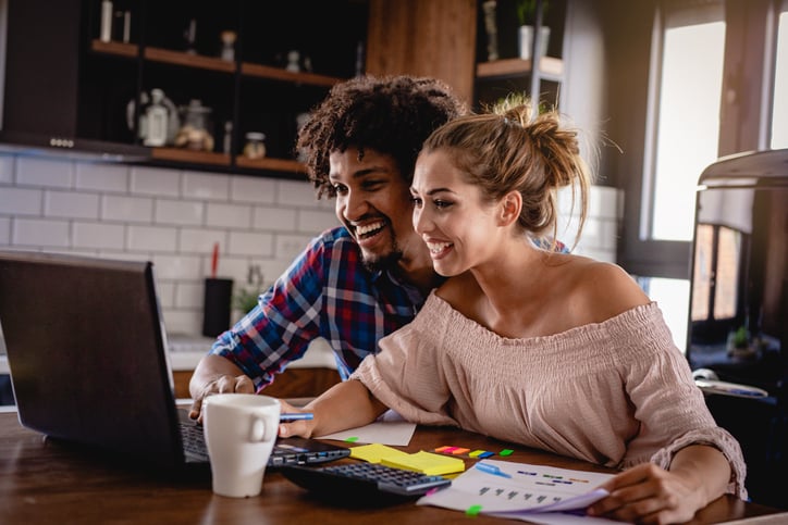 Happy couple checking their bank accounts on a laptop and paper at a desk