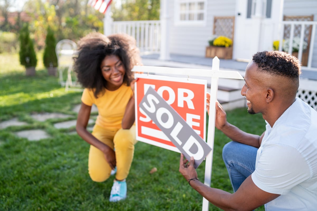 A young couple happily attaches a "Sold" banner to a "For Sale" sign outside a house.