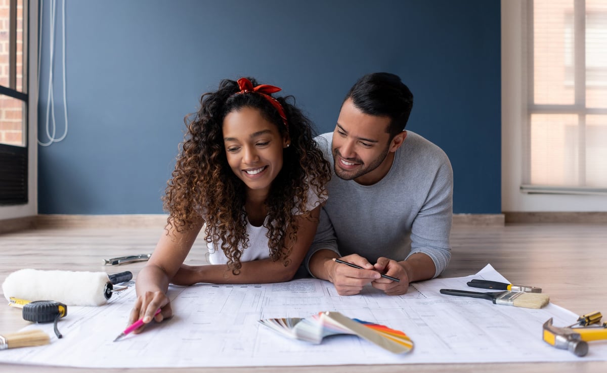 A couple looks at the blueprints of their new house while lying on the floor smiling.