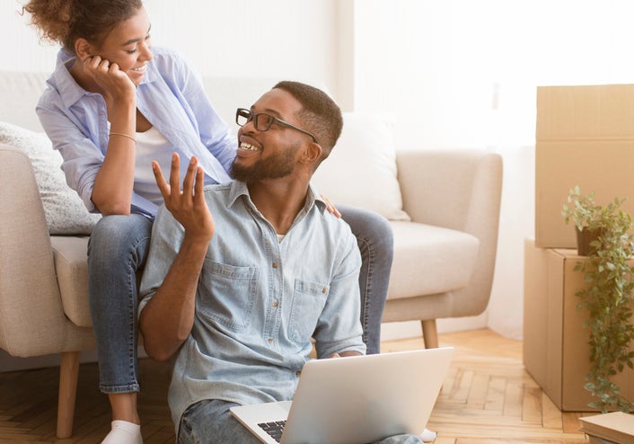 A couple sitting in their living room, smiling as they look at a laptop.