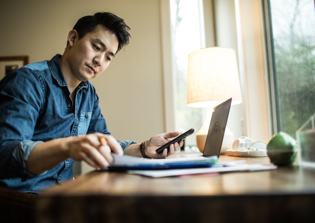 A person flips through papers at a home desk with a phone and laptop.