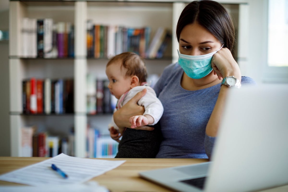 A worried caregiver wearing a face mask works from home while holding a baby.