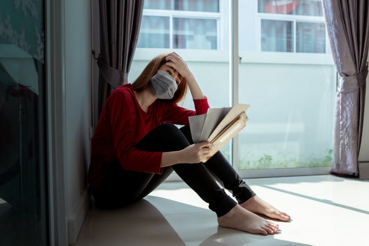 Masked woman sits on floor holding handful of bills in one hand and her head in the other.