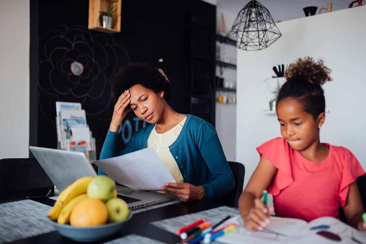 A worried caregiver works on a laptop next to their homeschooled child.