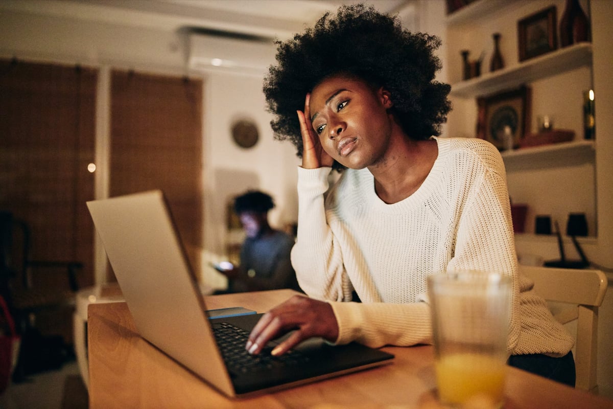 A person looks worriedly at their computer in their home, resting their hand on their head.