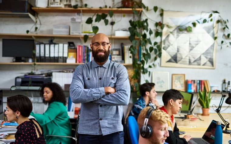 A bald person with a beard is standing in an office with creative colleagues and smiling at the camera.