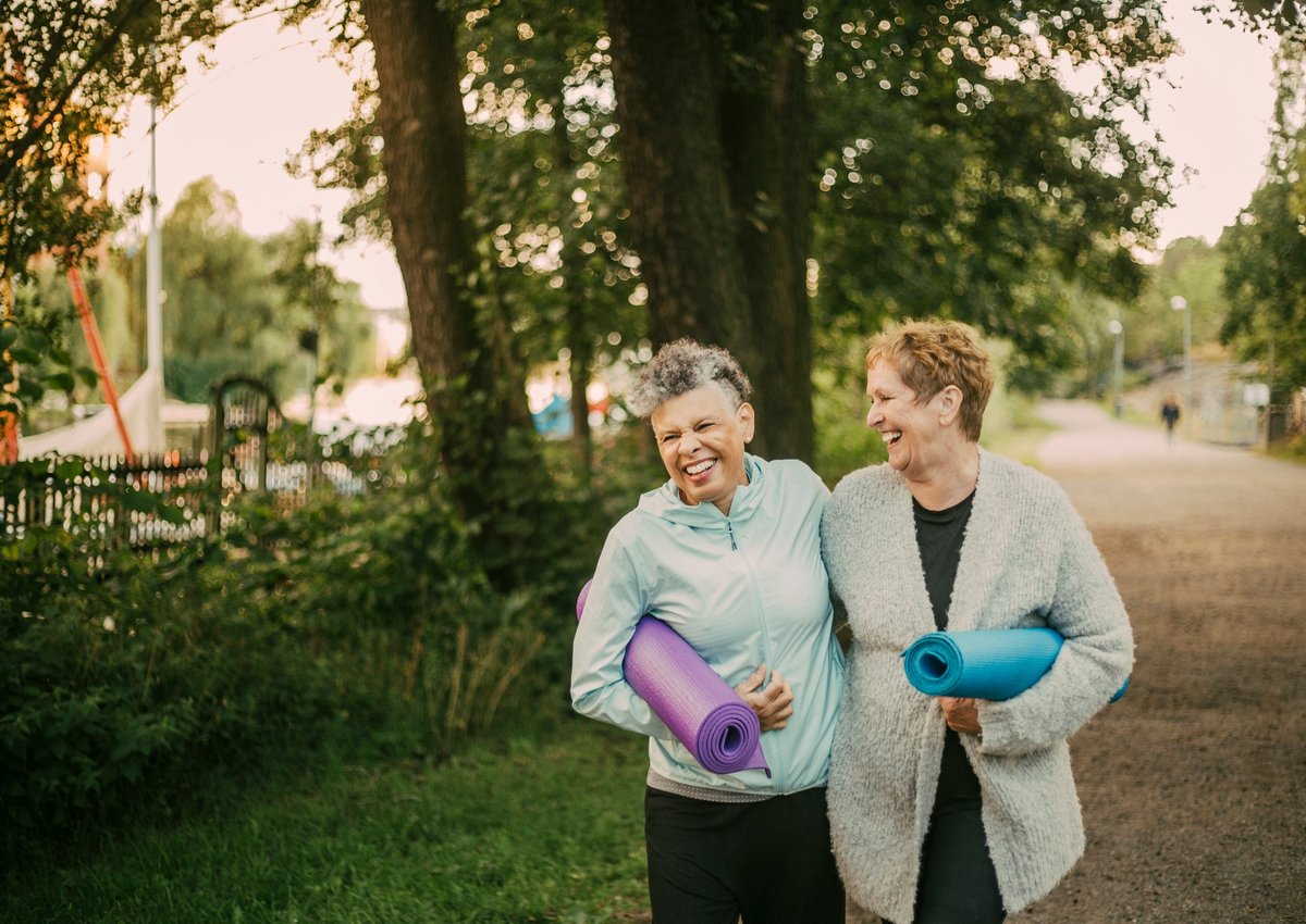 An older couple laugh with their arms around each other while holding yoga mats outside.